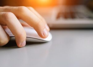 A close-up of a hand using a white computer mouse next to a laptop on a desk, with warm sunlight in the background.