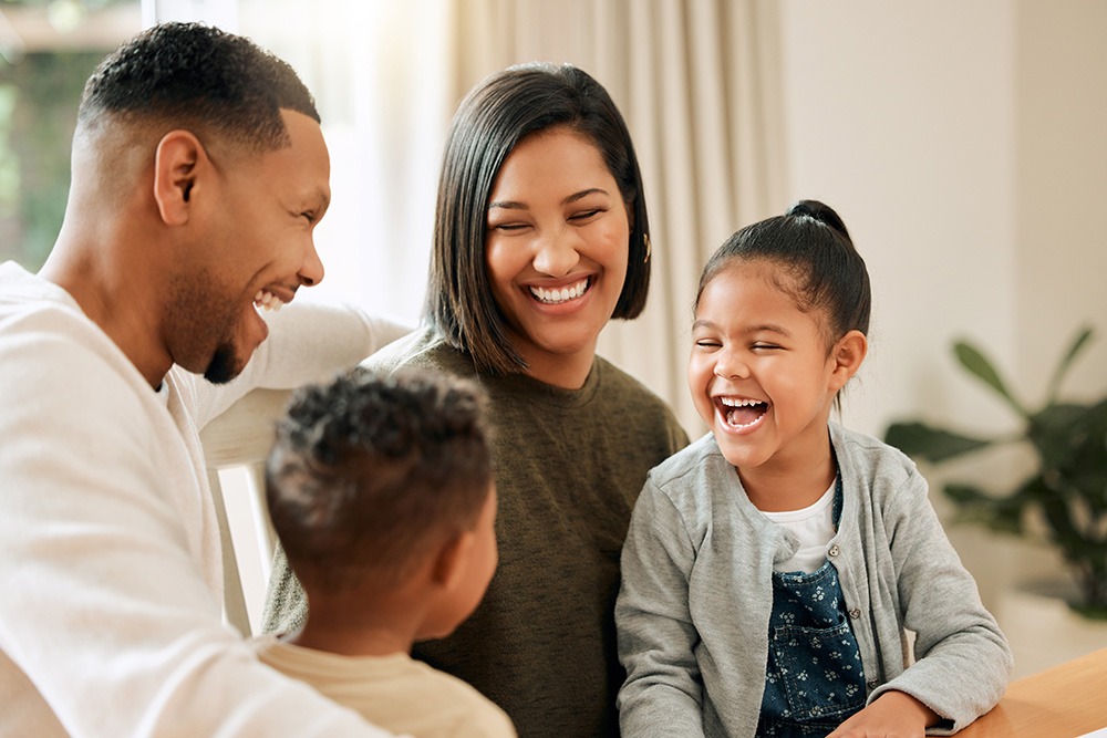 A smiling family of four, including two parents and two young children, sits together indoors, laughing and enjoying each others company.