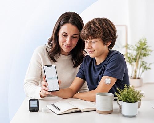 A woman and a boy with a glucose monitor on his arm look at a smartphone displaying a graph, reflecting their commitment to diabetes care Mishawaka IN. They sit at a table with a notebook, coffee mug, and plants.