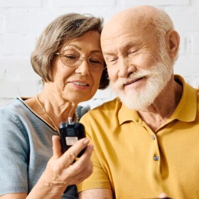 About: An elderly woman and man sit close together, smiling as they look at a medical device in the woman’s hand. About them, there is a sense of warmth and companionship—the man wears a yellow shirt, while the woman wears glasses and a light blue top.